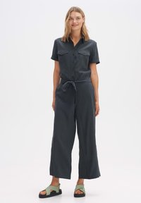 Dunkelgrüner Jumpsuit mit kurzen Ärmeln, Knopfverschluss vorne, zwei Brusttaschen und Taillengürtel. Kombiniert mit grünen und schwarzen Sandalen.