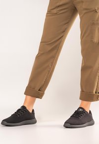 Schwarze Sportschuhe mit einer strukturierten Strickoberfläche, runder Spitze und kontrastierender grauer Sohle. Kombiniert mit hochgekrempelten beigen Hosen.