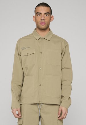 Tan button-up jacket with two chest pockets and prominent stitching. Features the text "HALO 19861972/1981" on the left side.