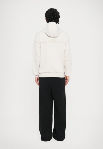 Ανοιχτό γκρι φούτερ με κουκούλα και άνετη εφαρμογή, με διακριτικές οριζόντιες ραφές και ριμπ μανσέτες, συνοδευόμενο από μαύρο παντελόνι με φαρδιά γραμμή.