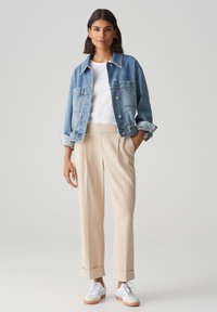 Giubbotto di jeans azzurro chiaro con due tasche frontali, abbinato a pantaloni beige con pieghe e sneakers bianche con suole in gomma.