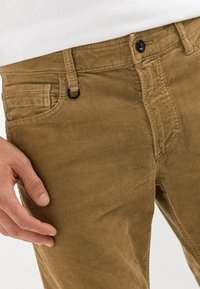 Pantalon en velours côtelé marron avec une texture nervurée, doté d'une fermeture à boutons et d'une poche latérale, main reposant sur le tissu.