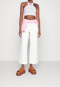 Kvinna som bär vita cropped byxor, en vit halterneck-top, en rosa sweatshirt knuten runt midjan, och bruna plattformssandaler.