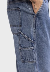 Personne portant un pantalon cargo bleu en denim avec plusieurs poches, avec une main partiellement dans la poche avant.