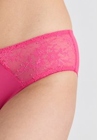 Rózsaszín csipkével hímzett bikini, szilárd anyag alapú. Átlátszó csipke díszítéssel az alsó él körül és ívelt lábnyílásokkal rendelkezik.