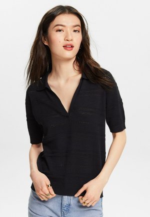 Giovane donna con capelli lunghi e scuri, che indossa un top a maglia nero a maniche corte con colletto e jeans di denim azzurro chiaro su uno sfondo bianco uniforme.