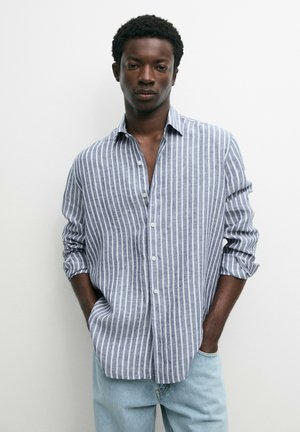 Man draagt een blauw-wit verticale gestreepte blouse met opgerolde mouwen, lichtblauwe spijkerbroek, staand tegen een effen witte achtergrond.