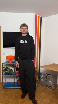 Mladý muž s tetovaniami stojí v rohovom priestore, oblečený v čiernej mikine značky Karl Lagerfeld a širokých čiernych nohaviciach, za ním je farebná pruhovaná stena.