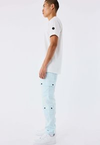 Uomo in piedi di lato, indossa una camicia bianca a maniche corte, pantaloni cargo azzurri chiari con bottoni neri e sneakers bianche, su uno sfondo bianco.