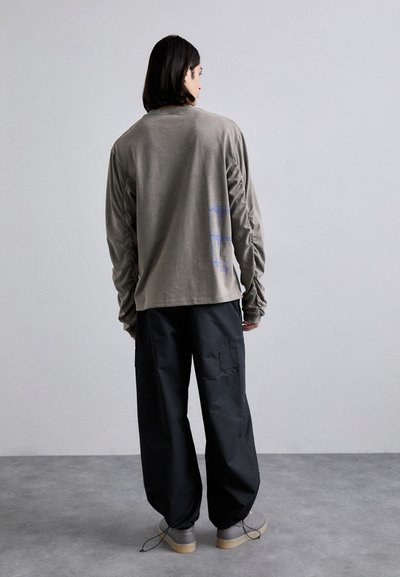 Camicia di cotone a maniche lunghe, verde oliva, con dettagli grafici blu, abbinata a pantaloni cargo neri e sneakers grigio chiaro su uno sfondo neutro.