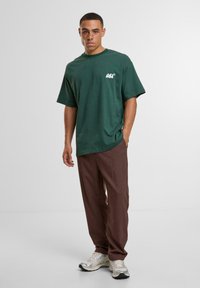 Green cotton t-shirt with a printed white logo on the left chest, paired with brown trousers and silver sneakers.