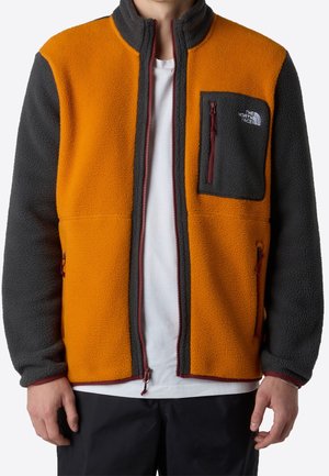 Mężczyzna w pomarańczowej i ciemnoszarej polarowej kurtce z kieszeniami na suwak i logo The North Face na kieszeni na piersi.