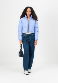 Kappet dunjakke i lys lilla med høy krage, glidelås og quiltet mønster, parret med vide blå jeans og hvite sko.
