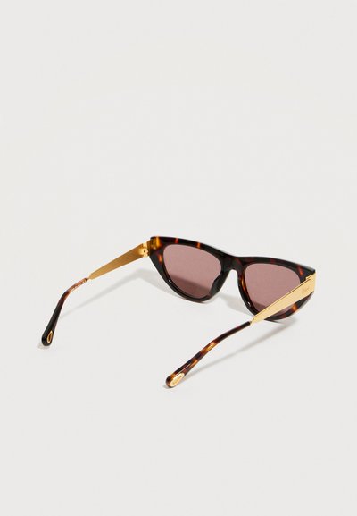 Γυαλιά ηλίου cat-eye με όψη χελώνας, σκοτεινούς φακούς και χρυσούς μεταλλικούς βραχίονες σε ανοιχτό φόντο.
