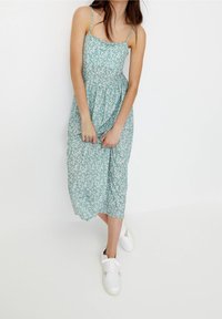 Femme portant une robe midi fleurie verte à fines bretelles et des baskets blanches, debout devant un fond blanc uni.