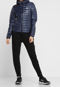 Veste matelassée bleu marine avec capuche, motif matelassé horizontal et fermeture éclair à l'avant. Portée avec un pantalon de jogging noir et des chaussures de sport noires.