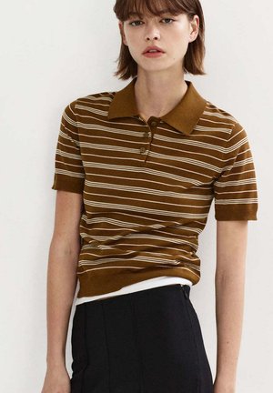 Jeune femme portant un polo à manches courtes rayé marron et blanc et un pantalon noir sur un fond clair uni.