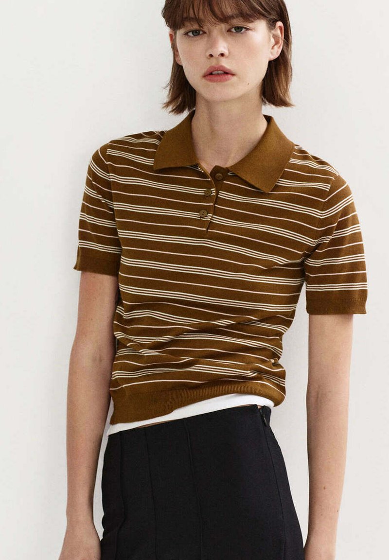 Jeune femme portant un polo à manches courtes rayé marron et blanc et un pantalon noir sur un fond clair uni.