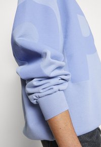 Pull bleu clair à manches longues avec poignets côtelés, porté par une personne à la peau claire avec des poils visibles sur l'avant-bras.