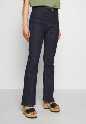 Personne portant un jean bootcut bleu foncé et des sandales plateformes noires avec talons en bois, debout devant un fond clair uni.