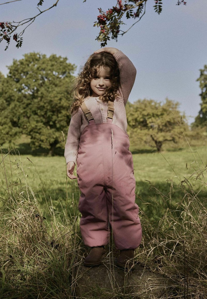 Salopette rose rembourrée avec des bretelles beiges ajustables, associée à un pull en tricot rose pâle. L'enfant se tient dans des herbes hautes à l'extérieur.