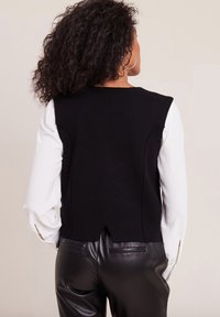 Gilet nero lavorato a maglia con un design corto, caratterizzato da un'apertura sul retro, abbinato a una camicia bianca a maniche lunghe e pantaloni di pelle neri.