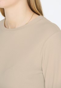Rippiges beiges Oberteil mit Rundhalsausschnitt, das eine glatte Textur und eine enganliegende Passform aufweist, betont durch dezente Strickmuster und saubere Kanten.