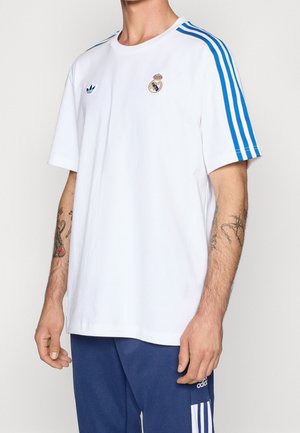 Mann trägt ein weißes Real Madrid Adidas-T-Shirt mit blauen Streifen an den Ärmeln, kombiniert mit blauen Adidas-Hosen mit weißen Streifen.