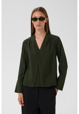 Femme portant un chemisier vert foncé à manches longues et col en V, un pantalon noir, une main dans la poche et de petites lunettes de soleil noires sur fond gris.