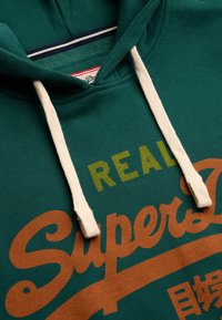 Sweat à capuche vert foncé avec cordons blancs et texte stylisé jaune et orange affichant « REAL SuperDry » avec des caractères japonais partiellement visibles.