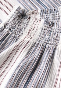 Witte stof met blauwe en rode verticale strepen, met een textuur van smocking en gerimpelde details langs de bovenrand.