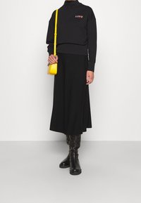 Sweat-shirt noir avec broderie "LOVE", associé à une longue jupe noire et des bottes noires montantes. Un sac bandoulière jaune apporte du contraste.