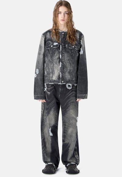 Jeune personne aux longs cheveux ondulés portant une veste en jean noire et grise délavée surdimensionnée et un jean ample assorti, debout devant un fond blanc.