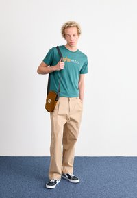 Camiseta de manga corta color teal con el logo "ALPHA INDUSTRIES", pantalones de pierna ancha beige, bolso cruzado marrón y zapatillas negras con suelas blancas.