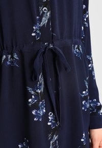 Abito button-up floreale blu navy con maniche lunghe, vita regolabile con coulisse e tessuto testurizzato che presenta motivi floreali blu chiaro e bianchi.