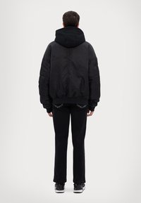Giubbotto bomber nero con cappuccio, caratterizzato da una texture liscia e polsi elasticizzati, abbinato a pantaloni neri e scarpe da ginnastica. Visualizzazione della parte posteriore.
