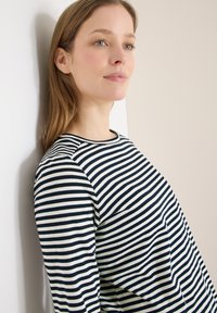 Jeune femme aux cheveux châtain clair portant un haut à manches longues rayé noir et blanc, appuyée contre un mur blanc et beige.