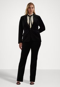 Costum negru din catifea cu un sacou ajustat și pantaloni drepți. Bluză crem cu volane și cravată neagră. Sandale negre simple.