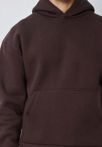 Sweat-shirt à capuche marron avec une poche kangourou, fabriqué en tissu doux. Présente une coupe décontractée et un design simple et épuré.