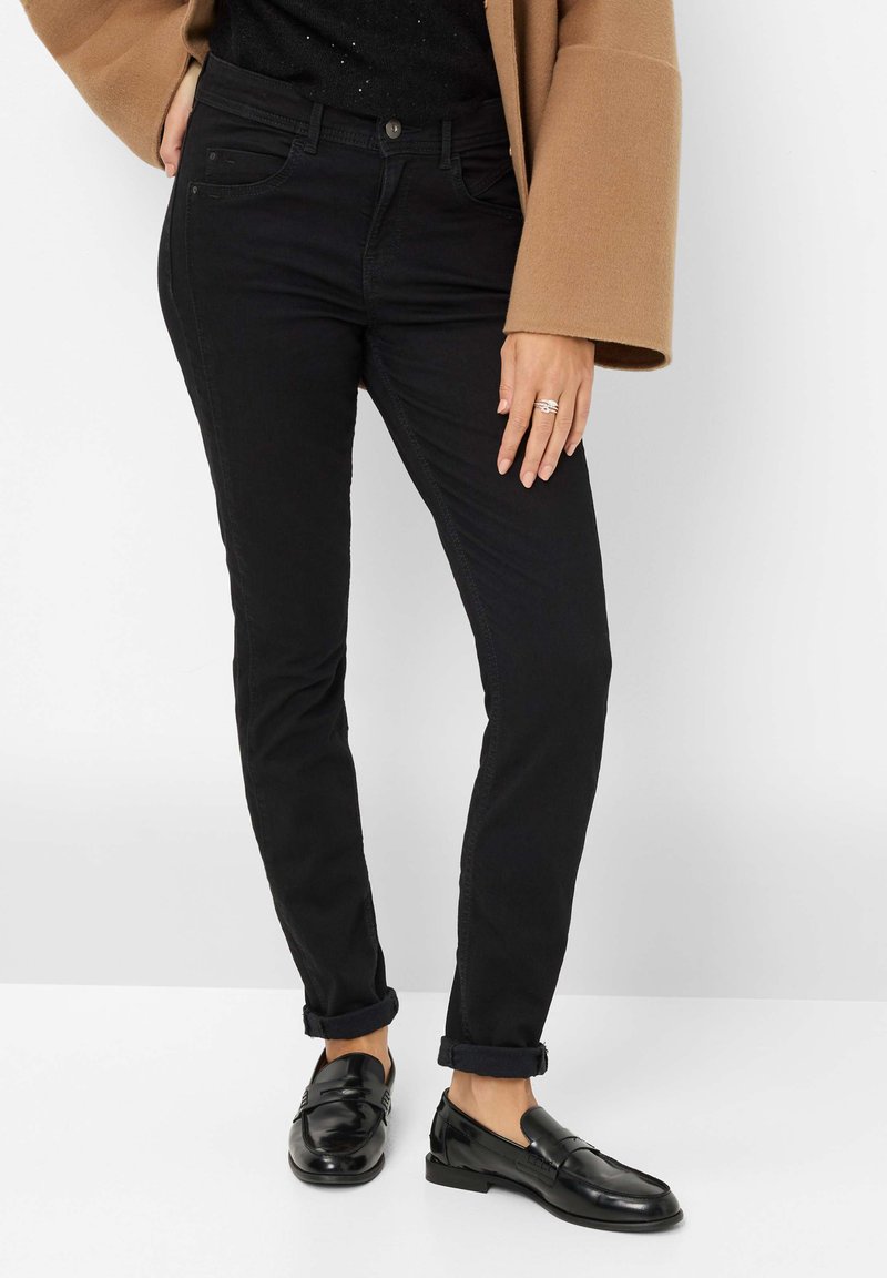 Jean noir moulant avec une légère élasticité, taille mi-haute et ourlets repliés, associés à des mocassins noirs brillants. Tissu lisse et texturé.