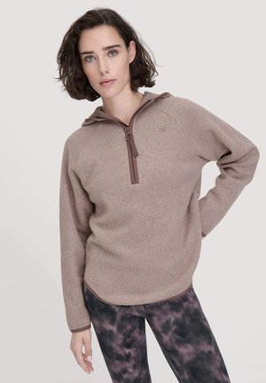 Pullover van zachte, beige fleece met een half-rits kraag en een bruine rits. Lange mouwen met contrasterende boorden. Gekleed met tie-dye leggings.