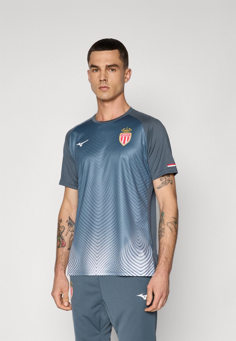 Junger Mann mit kurzem dunklem Haar, der ein grau verlaufendes AS Monaco Sporttrikot und passende Hose trägt, steht vor einem schlichten Hintergrund.