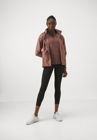 Giacca leggera con cappuccio in rosa polveroso sopra una maglietta bordeaux, abbinata a leggings neri e sneakers bianche. Tessuto liscio, vestibilità atletica.