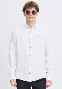 Camisa de lino blanca de manga larga con botones en el frente, que presenta un pequeño logo negro en el pecho y un dobladillo curvado.