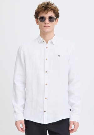 Chemise blanche en lin à manches longues, avec des boutons sur le devant, affichant un petit logo noir sur la poitrine et un ourlet arrondi.