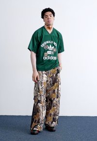 Zöld, oversized Adidas mez emblémákkal, laza, katonai mintás cargo nadrággal párosítva. Fekete cipők. Egyszerű háttér és kék szőnyeg.