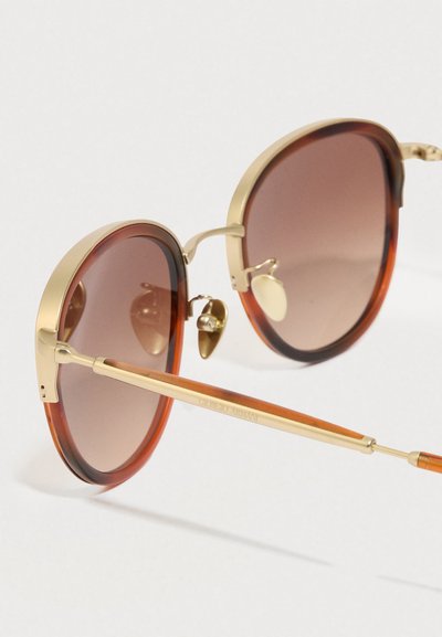 Kerek napszemüveg teknőcskagyló mintás kerettel és aranyozott díszítéssel. Barna színátmenetes lencsék. Fém orrnyereg és állítható orrtámaszok a kényelemért.