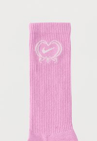 Roza atletična nogavica z rebrasto teksturo, na kateri je bel logotip srca z Nikeovým kljukastim simbolom in dva dekorativna loka na podnožju.