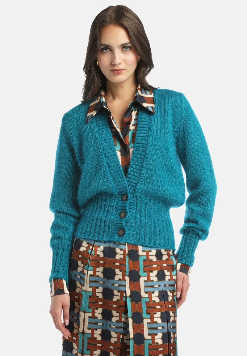 Türkiser Wollcardigan mit V-Ausschnitt, geripptem Saum und drei braunen Knöpfen, kombiniert mit einem gemusterten braunen und türkisfarbenen Rock.