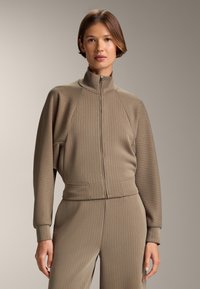 Hnedá rebrovaná bunda na zips s vysokým golierom, ktorá má raglánové rukávy a predné vrecká. Kombinovaná s vhodnými rebrovanými nohavicami.